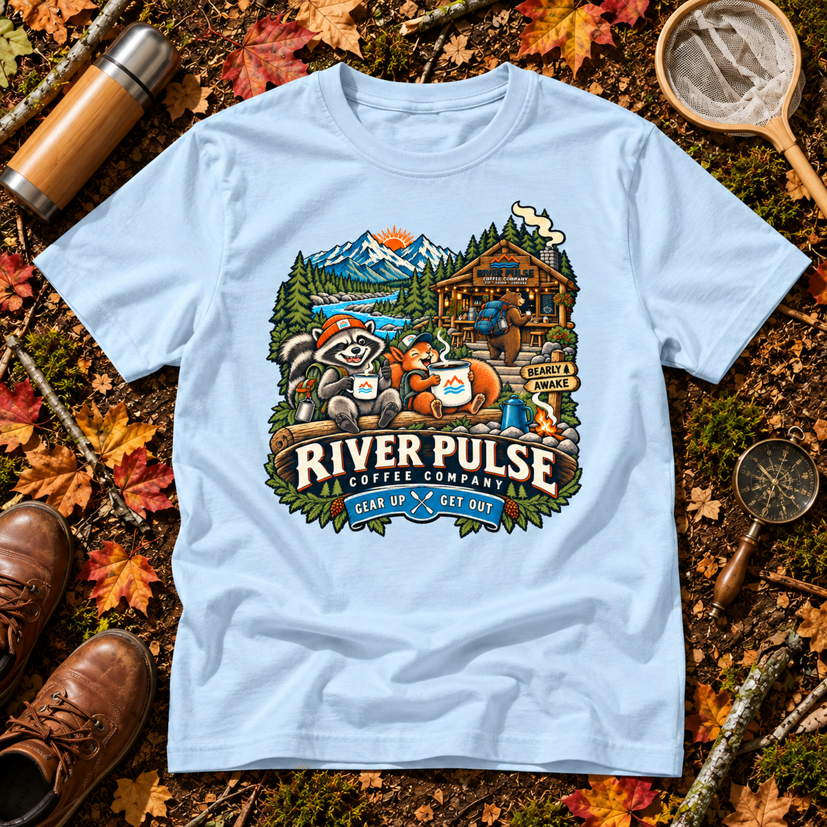 A light blue graphic t-shirt laid flat on a forest floor surrounded by autumn leaves, a thermos, a magnifying glass, and brown boots. The t-shirt features a detailed graphic of raccoons, a cabin, mountains, and the text 'RIVER PULSE COFFEE COMPANY GEAR UP & GET OUT'.