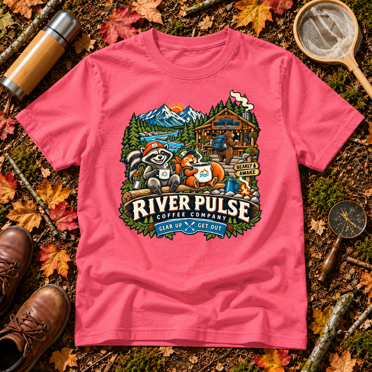 A pink short-sleeve graphic T-shirt laid flat on a forest floor with autumn leaves, boots, a thermos, and a magnifying glass. The shirt features a large illustrated front print with a raccoon, fox, mountain cabin, trees, and the text 'RIVER PULSE CLOTHING COMPANY' and 'GEAR UP & SEE OUT'.